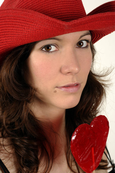 Image of a beautiful young woman wearing a red cowboy and holding a heart-shaped lollipop. - Fotoğraf, Görsel