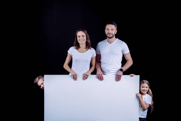 Smiling family standing with blank white card and looking at camera on black - Foto, Imagem