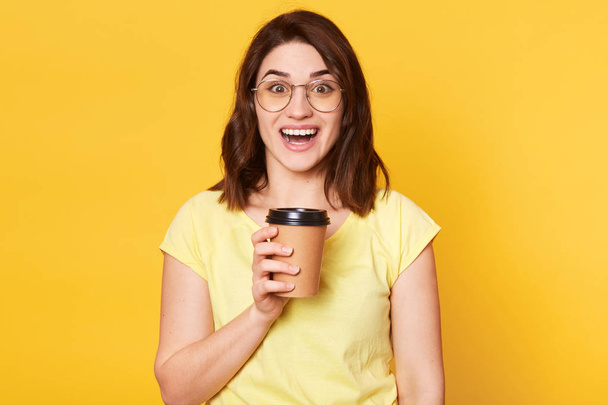 Close up portrait of young lady isolated on yellow studio background, posing with happy facial expression while drinking tekeaway coffee, has positive feelings and toothy smile, stands with open mouth - Foto, imagen