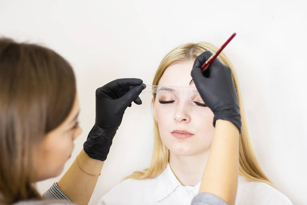 Female cosmetologist performs eyebrow correction on beautiful models in the beauty parlor. The girl's face is a blonde. Close-up. Facial care - Zdjęcie, obraz