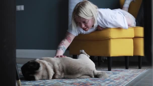 Young woman pet her dog sitting on the chair. Cute puppy pug sleeping on the floor. Scene of love and care. - Video