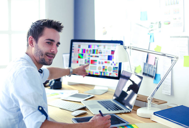 Portret młodego projektanta siedzącego w pracowni graficznej przed laptopem i komputerem podczas pracy online. - Zdjęcie, obraz
