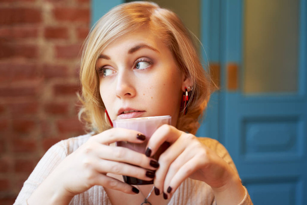 A beautiful woman with short blond hair and blue eyes looks out the window. Successful woman thinking about work, holding a Cup of coffee. Large picture of the person in the cafe - Foto, Imagen