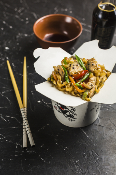 Fideos chinos en una caja con pollo y verduras con palitos. Wok entrega de comida del restaurante sobre fondo oscuro desde la vista lateral
. - Foto, imagen