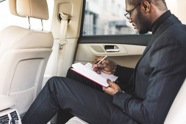 A young business man in a suit is sitting in the back seat of a expensive car with a notebook. Business negotiations. - Fotografie, Obrázek