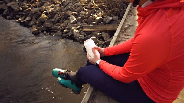 Caucasian woman sportsman in bright sports shoes helmet rests on the bridge cross the river. Cyclist in bright sportswear with a flask a plastic bottle in his hand. Rest stop break drink water thirst - Photo, Image