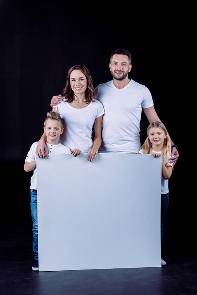 Smiling family in white t-shirts holding blank white card and looking at camera - Foto, immagini