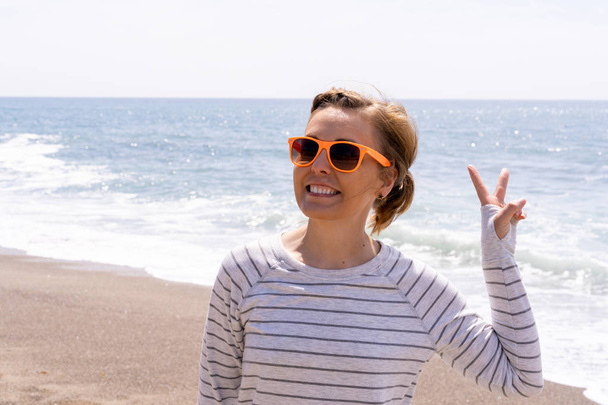 Caucasian woman on the beach gives a peace sign, wearing casual  - Photo, Image