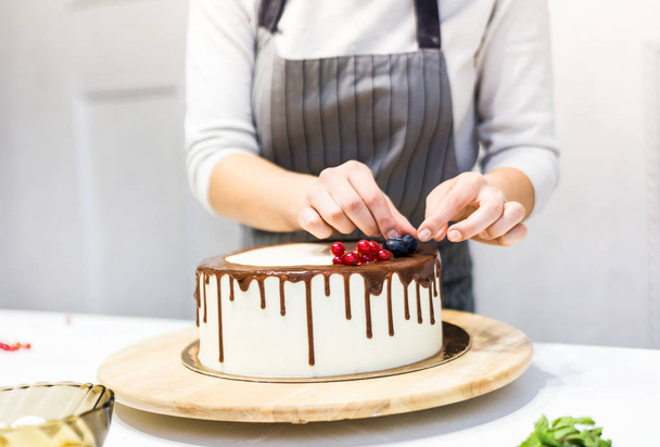 Confectioner decorates with berries a biscuit cake with white cream and chocolate. Cake stands on a wooden stand on a white table. The concept of homemade pastry, cooking cakes. - 写真・画像