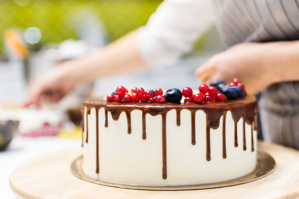 Confectioner decorates with berries a biscuit cake with white cream and chocolate. Cake stands on a wooden stand on a white table. The concept of homemade pastry, cooking cakes. - Photo, Image