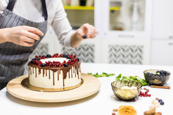 Confectioner decorates with berries a biscuit cake with white cream and chocolate. Cake stands on a wooden stand on a white table. The concept of homemade pastry, cooking cakes. - Фото, изображение