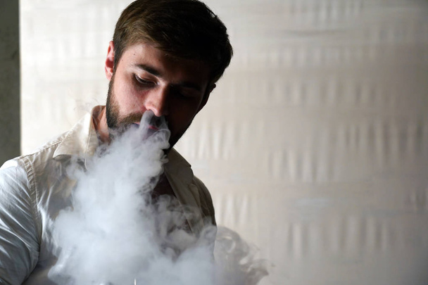 Πορτρέτο ενός βίαιου μουσάτο καυκάσιο άντρα με κοντό καστανά μαλλιά σε λευκό πουκάμισο σε θολό φόντο. Ο όμορφος άντρας καπνίζει ένα τσιγάρο και εκπνέει ένα σύννεφο καπνού από το στόμα του με ευχαρίστηση.. - Φωτογραφία, εικόνα
