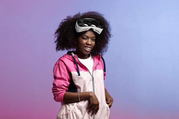 Funny curly brown-haired girl dressed in the pink sports jacket is wearing on her head the virtual reality glasses in the studio on neon background - Foto, Bild