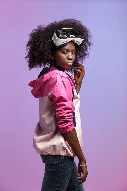 Mod curly brown-haired girl dressed in the pink sports jacket wearing on her head the virtual reality glasses poses in the studio on neon background - Fotoğraf, Görsel