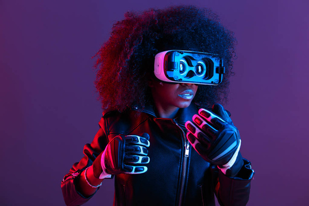 Curly dark haired girl dressed in black leather jacket and gloves uses the virtual reality glasses on her head in the dark studio with neon light - Foto, Imagem