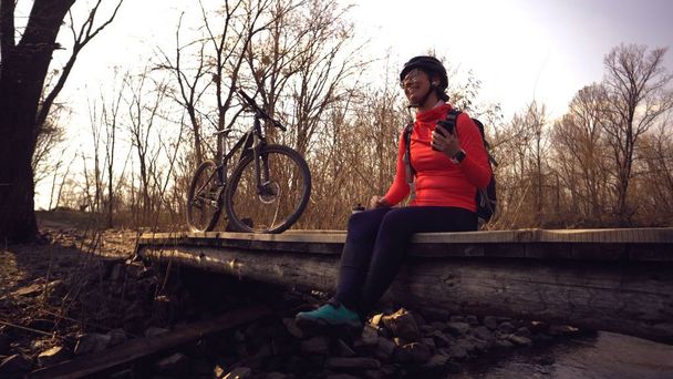 Happy young woman sitting wooden bridge over river, listening music in headphones with smartphone hands and dancing. Positive cyclist listens song in nature. Athlete happy near bike ears earphones - Photo, Image