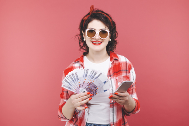 Πορτραίτο ενός χαμογελαστή κοριτσιού, κρατώντας ένα μάτσο χαρτονομίσματα και κοιτάζοντας το κινητό τηλέφωνο απομονωμένη πάνω από ροζ - Φωτογραφία, εικόνα