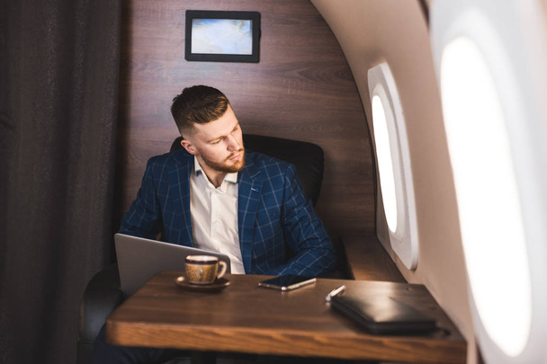 Attractive and successful businessman working on a laptop while sitting in a chair of his private jet. - Foto, Imagem