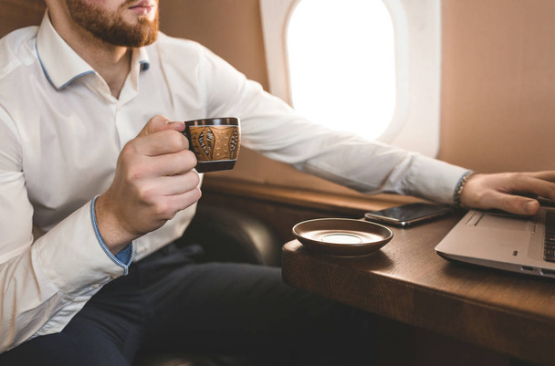 Atractivo y exitoso hombre de negocios bebiendo café y trabajando detrás de un portátil mientras está sentado en una silla de su jet privado
. - Foto, Imagen