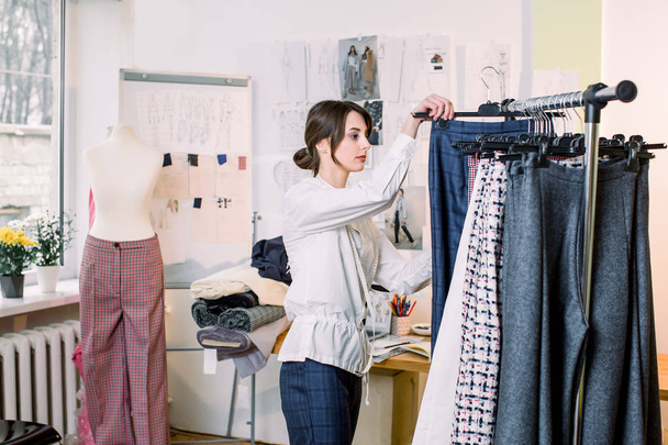 Dressmaker, tailor or needlewoman standing near clothing rack with fashionable stylish handmade clothes pants. Successful fashion designer. - 写真・画像