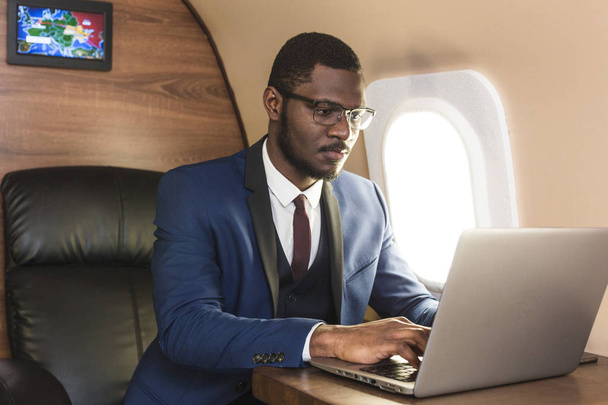 Attractive and successful African American businessman with glasses working on a laptop while sitting in the chair of his private jet. - 写真・画像