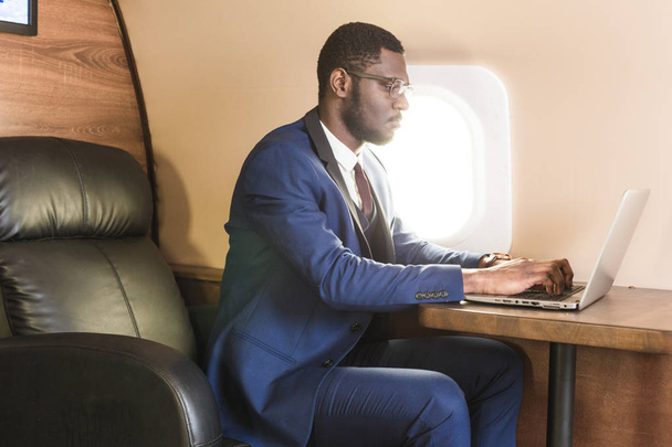 Attractive and successful African American businessman with glasses working on a laptop while sitting in the chair of his private jet. - Φωτογραφία, εικόνα