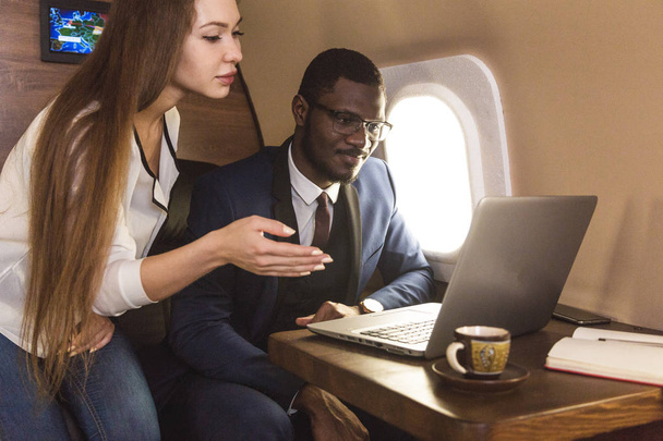 Young successful African-American businessman with glasses and an attractive female workmate blonde in a private jet. Service and flight first class - Фото, изображение
