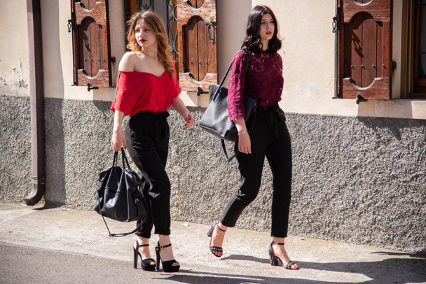 Két női barát sétál az utcán. A szőke egy piros inget és fekete nadrágot, a másik barna egy piros csipke ing és egy fekete melltartó fekete nadrágot. A háttérben az ajtó és ablak egy ház. - Fotó, kép