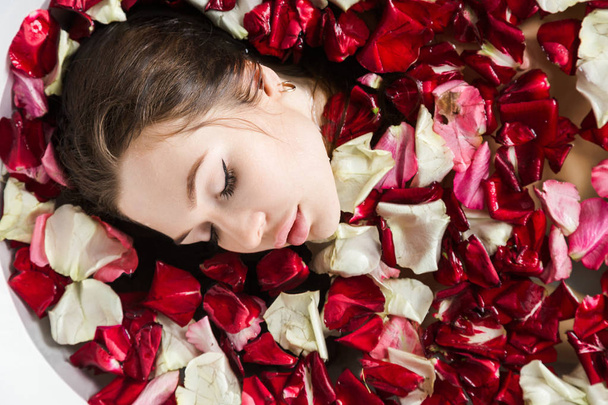 A young attractive girl takes a bathroom with flower petals and relaxes against the background of a beautiful light interior. Spa treatments for beauty and health with skin care - Fotografie, Obrázek