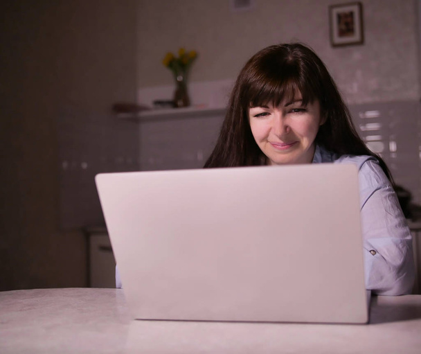 niedlich lächelnde junge brünette Frau sitzt am Abend in ihrer Küche mit einem Laptop - Foto, Bild