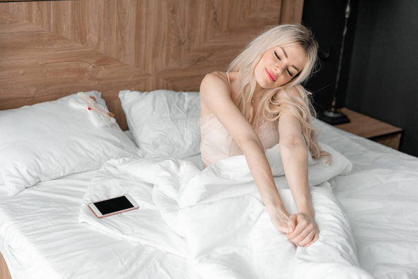 Morning in hotel room. Young woman wake up, sits on comfortable bed. Wooden headboard on background. - Foto, afbeelding