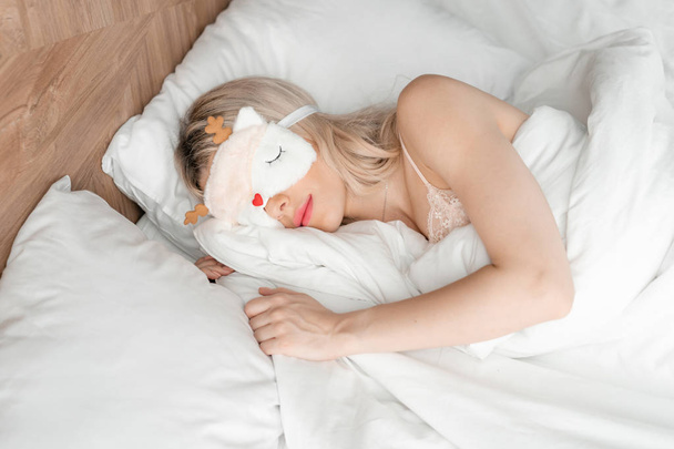 Mladá žena spí na pohodlné posteli v masce na spaní. Zavázané oči. Dobré ráno v hotelovém pokoji. Bílý polštář a přikrývka - Fotografie, Obrázek