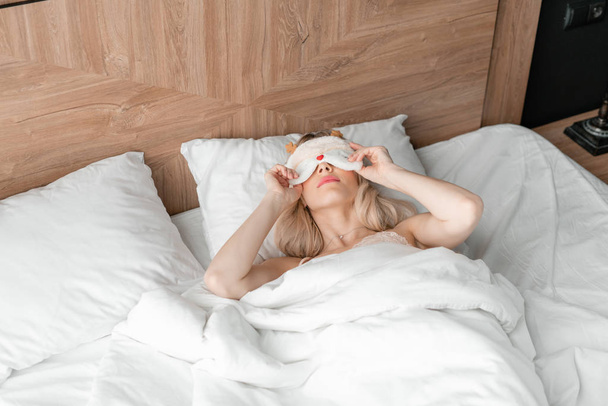 Genç bir kadın uyumak için rahat bir yatakta maske takar. Gözlerim bağlıydı. Otel odasında sabah oldu. Beyaz yastık ve battaniye - Fotoğraf, Görsel