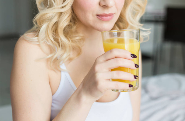 Attractive blonde woman sits on the edge of the bed at home and holds a stocker with orange juice in her hand. Morning mood and vigor - 写真・画像
