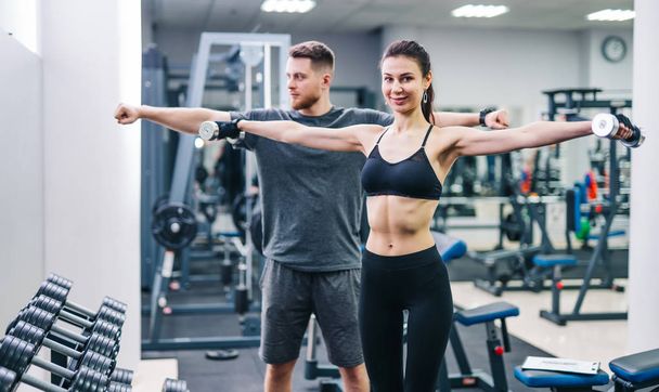 Smiling female in black top and leggings holding dumbbells before her coach at gym. Male trainer and beautiful woman lifting metal dumbbells and looking at camera. - Photo, Image