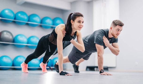 Young couple doing push-ups from the floor on one hand together on the background of fitness balls. Sporty male and female in sportswear training in the sports club. - Zdjęcie, obraz