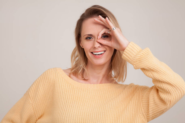 close up portrait of smiling blonde woman with white teeth, looking at the camera through fingers in ok gesture. wearing yellow sweater. poses against grey background. face expression, emotions - Photo, Image