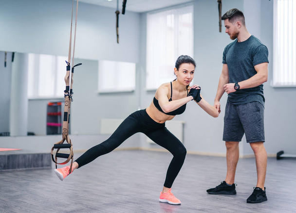Slim female doing legs training on TRX with the help of her personal trainer in fitness club. Young woman doing suspension rope exercise on leg holding balance next to the coach. - Zdjęcie, obraz