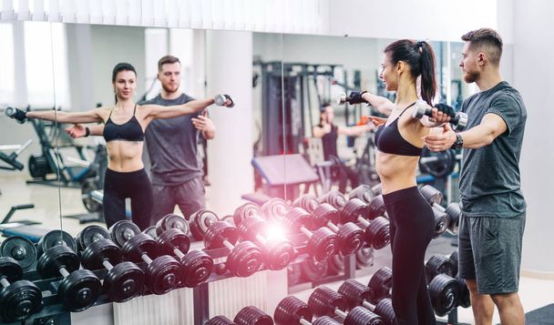 Slim sexy girl lifting dumbbells and male coach helping her in front of the mirror. Young woman and her trainer on the background of black dumbbells in a row. - Φωτογραφία, εικόνα