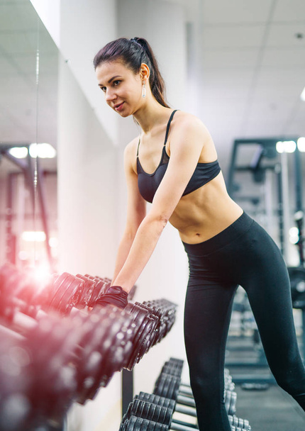 Attractive young woman taking dumbbells in the gym. Slim girl in black top and leggings training with heavy dumbbells. Close-up - Photo, Image