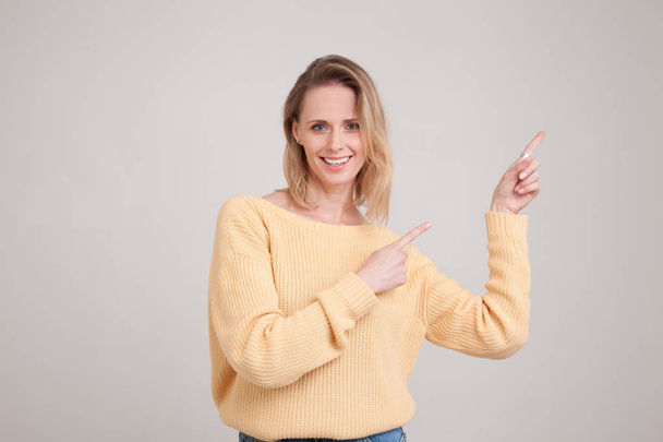 waist-up portrait of happy smiling blonde woman pointing fingers away, showing something interesting and exiting, looking at the camera. wearing yellow sweater. poses against grey background. face exp - Foto, Imagen