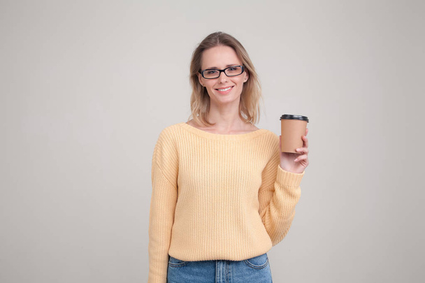Das hüfthohe Porträt einer blonden Frau, die eine Kaffeemütze in der Hand hält und in die Kamera lächelt. mit gelbem Pullover und Brille. posiert vor grauem Hintergrund. Gesichtsausdruck, Emotionen. - Foto, Bild