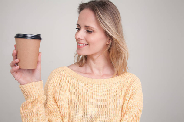 Porträt einer blonden Frau, die eine Kaffeemütze in der Hand hält, sie ansieht und lächelt. mit gelbem Pullover. posiert vor grauem Hintergrund. Gesichtsausdruck, Emotionen. - Foto, Bild