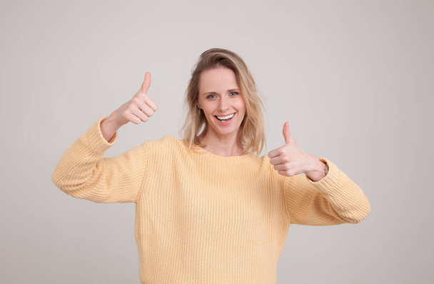 waist-up portrait go young blonde woman smiling and making thumbs up sign. showing respect to someone. i like it, good job. wearing yellow sweater. poses against grey background. face expression, emot - Photo, Image