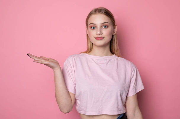 jong mooi blond meisje met blauwe ogen, het dragen van roze t-shirt tonen iets groot met open palm - Foto, afbeelding