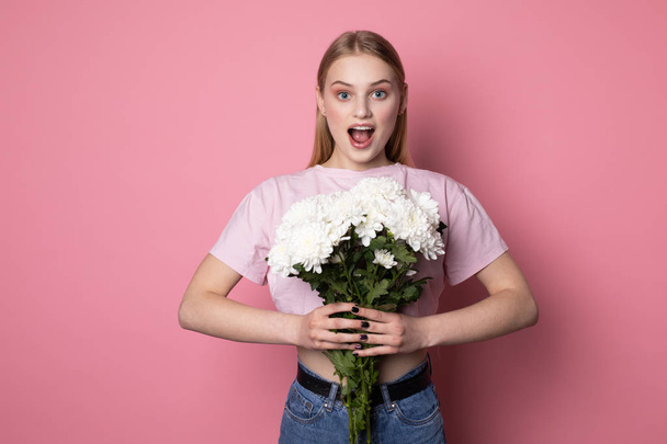 Young surprised blonde woman with open mouth wearing pink t-shirt with white flowers in hands - Foto, imagen