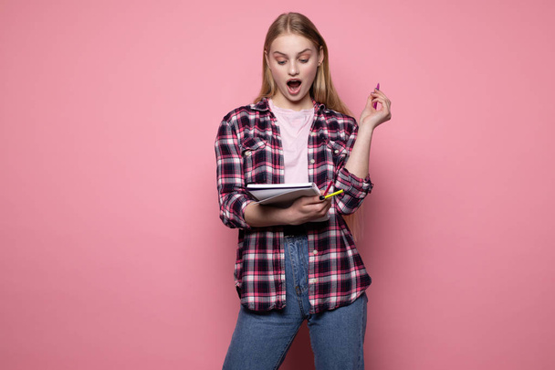 Young beautiful blonde girl in plaid shirt with open mouth showing eureka sign with her pen - Foto, afbeelding