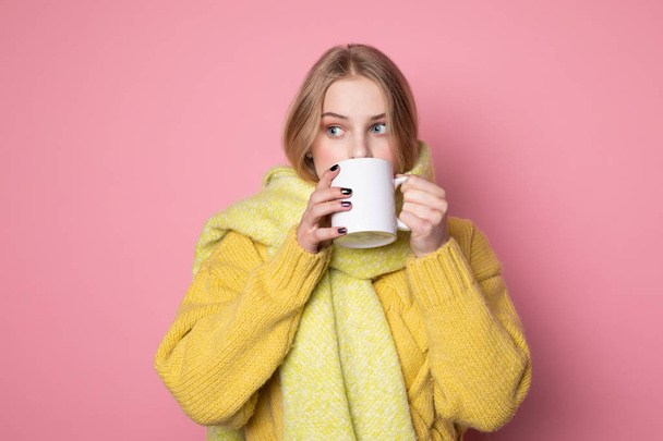 Scared blonde girl in yellow sweater and scarf, drinking hot tea - Fotografie, Obrázek