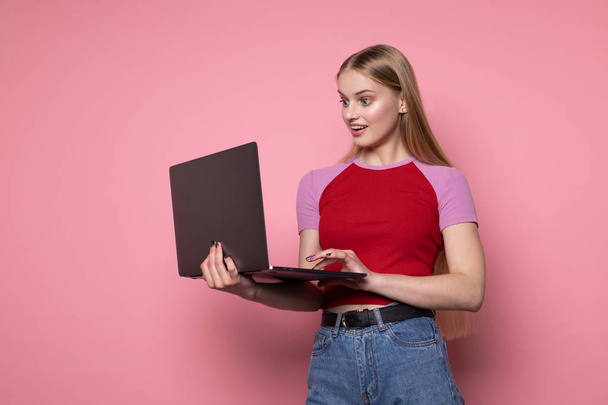 Happy smiling cheerful female with blond hair, wearing casual clothes, holding laptop in hand - Fotó, kép