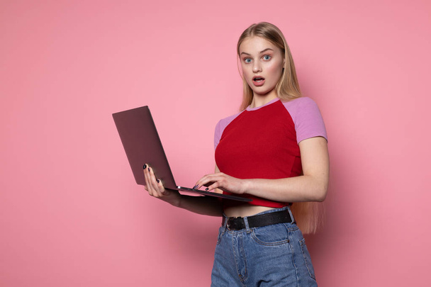 Портрет удивленной красивой девушки в красной футболке и синих джинсах с помощью ноутбука
 - Фото, изображение